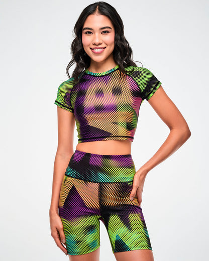 Female model wearing a purple, green, and tan crop top and matching biker shorts in a blurred, oversized "ZUMBA" wordmark print.