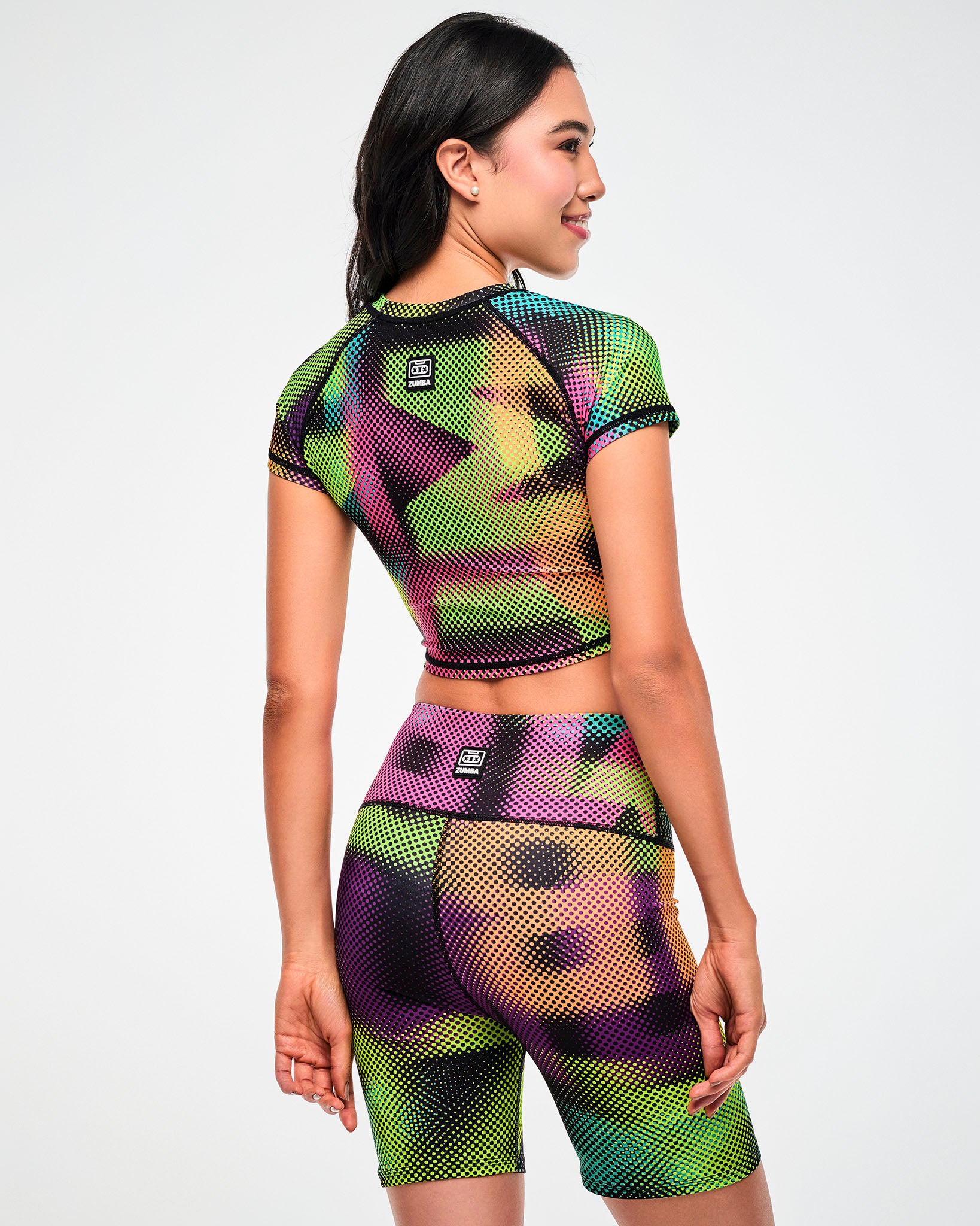 Female model wearing a purple, green, and tan crop top and matching biker shorts in a blurred, oversized "ZUMBA" wordmark print, shown from the back.