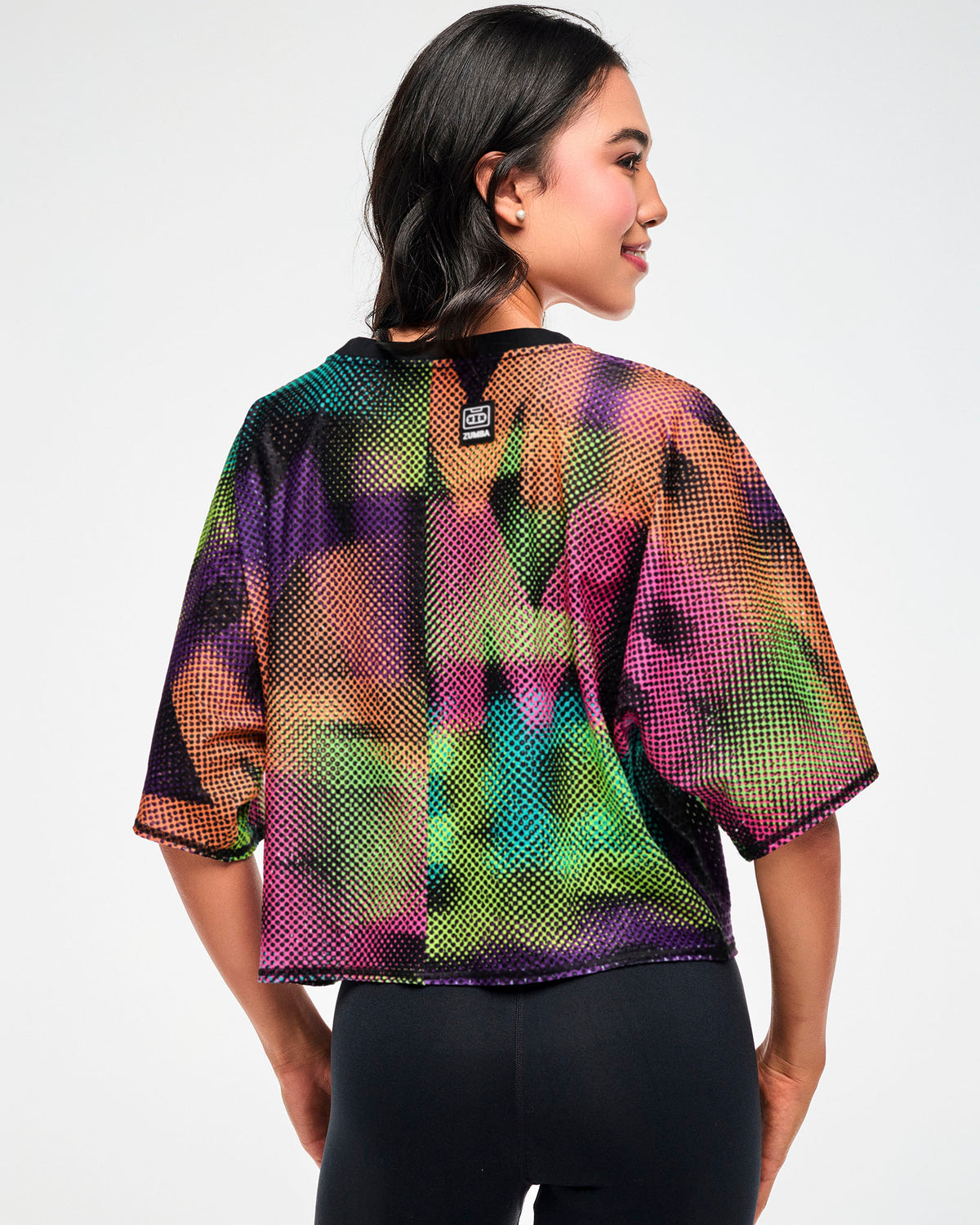 Shown from the back, female model wearing an oversized crop top with a blurred ZUMBA wordmark print, paired with black leggings.