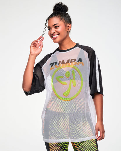 Female model wearing a white mesh oversized tee with black and white striped sleeves, a big yellow and orange Zumbito logo and "ZUMBA" text in black and orange above it, all paired with black and yellow dot-print leggings.