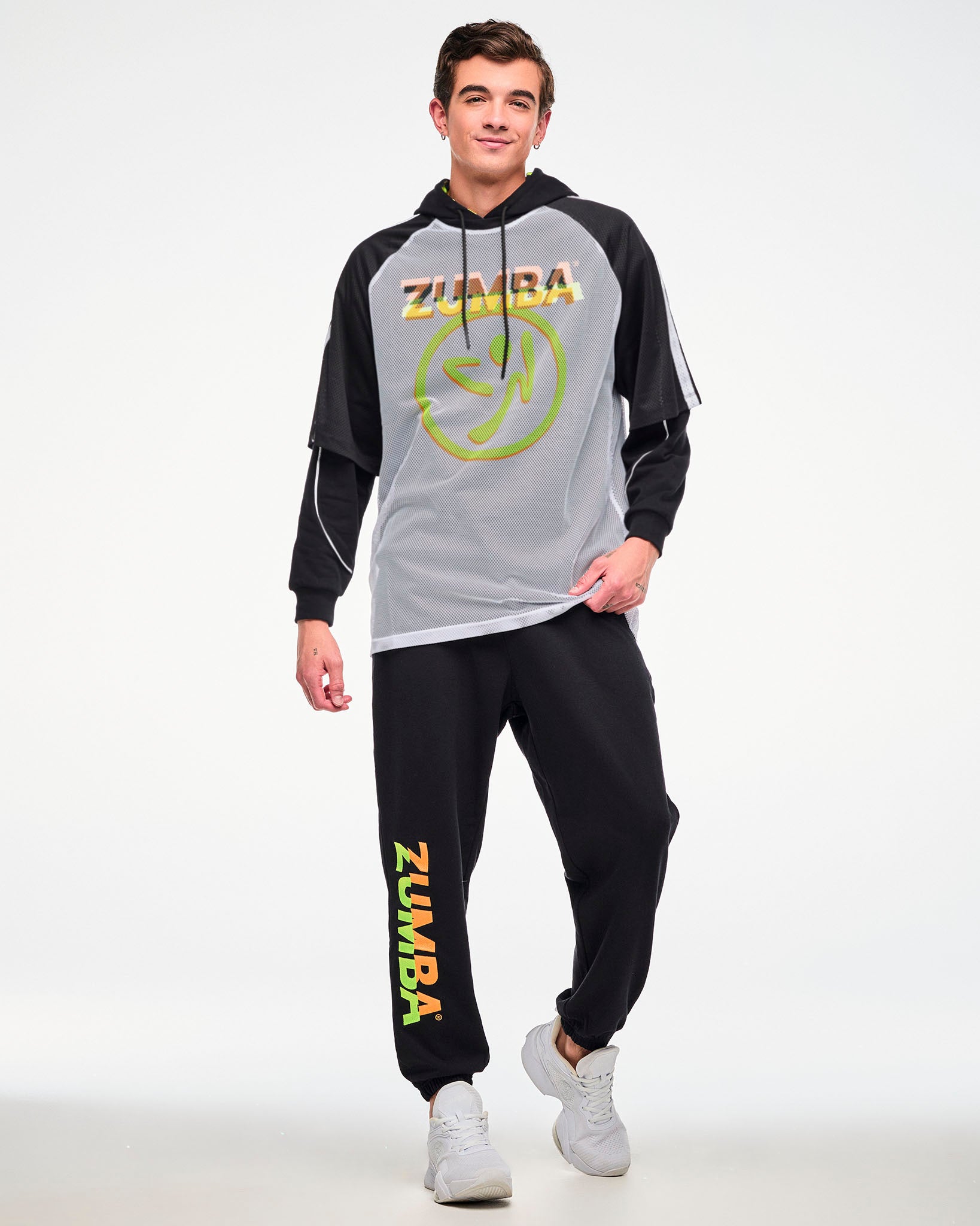 Male model wearing a white mesh tee with a big yellow and orange Zumbito logo and "ZUMBA" text in black and orange above it, paired with a black hoodie underneath, black sweatpants with Zumba text in orange and green and white shoes.
