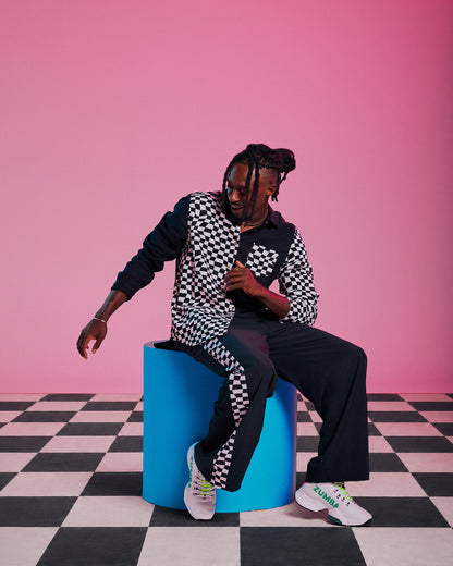 Male model wearing a black and white checkered flannel button-up, paired with black pants and white shoes, seated on a blue cylindrical pedestal against a solid pink background with a black and white checkered floor.