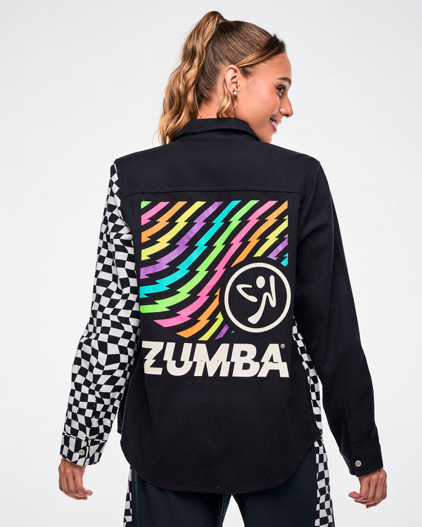 Shown from the back, female model wearing a black flannel button-up with a checkered sleeve and a rainbow lightning bolt Zumbito graphic and white 'ZUMBA' text on the back.