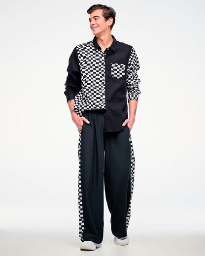 Full view of a male model wearing a black and white checkered flannel button-up, paired with black pants featuring a checkered side stripe and white shoes.