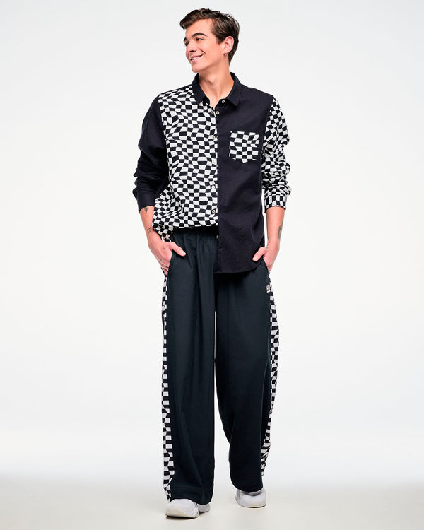 Full view of a male model wearing a black and white checkered flannel button-up, paired with black pants featuring a checkered side stripe and white shoes.