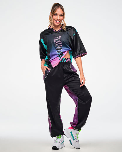 Female model wearing a black jersey tee with a pink-to-purple gradient "ZUMBA" print, paired with black joggers and white sneakers.