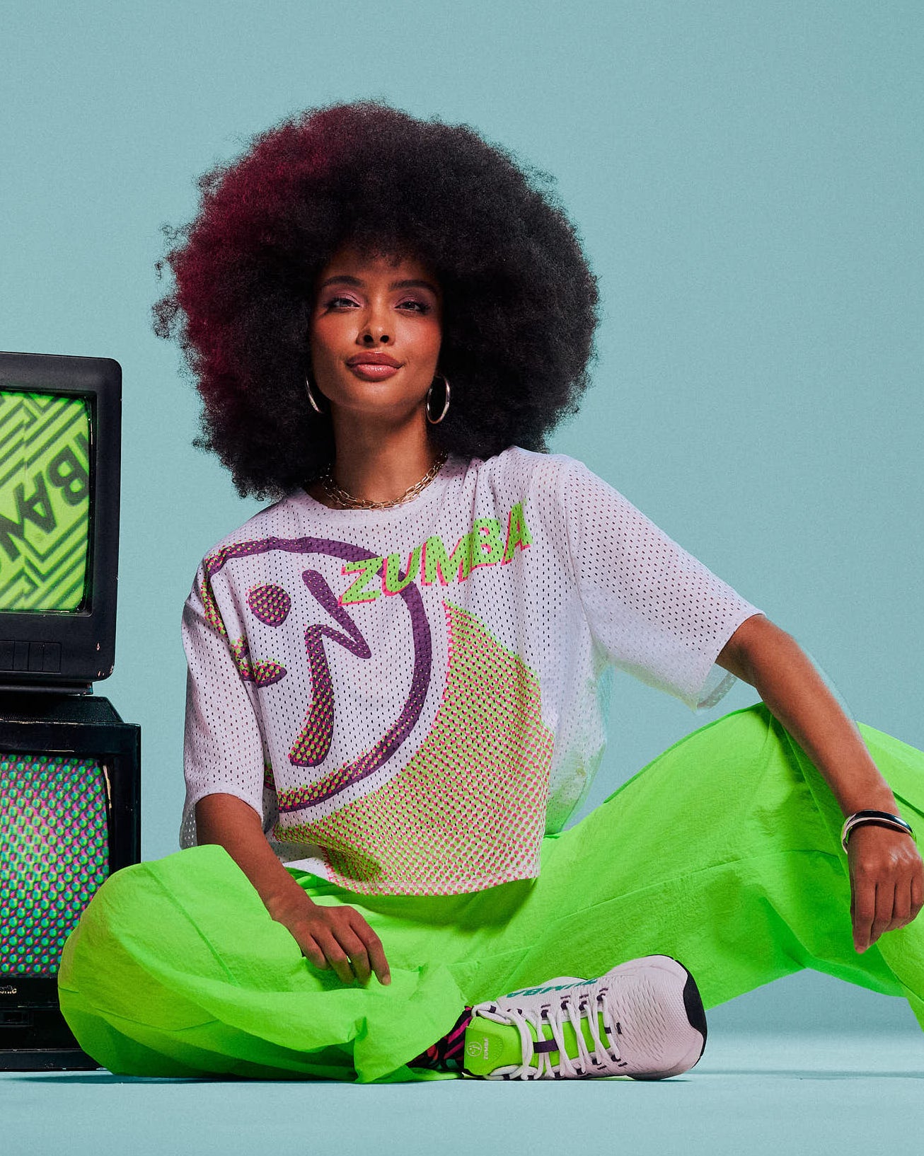 A woman with an afro sits on a light blue studio floor, next to 80's style TV's, wearing a white mesh crop top with a large purple Zumbito, paired with bright neon green pants and white sneakers.