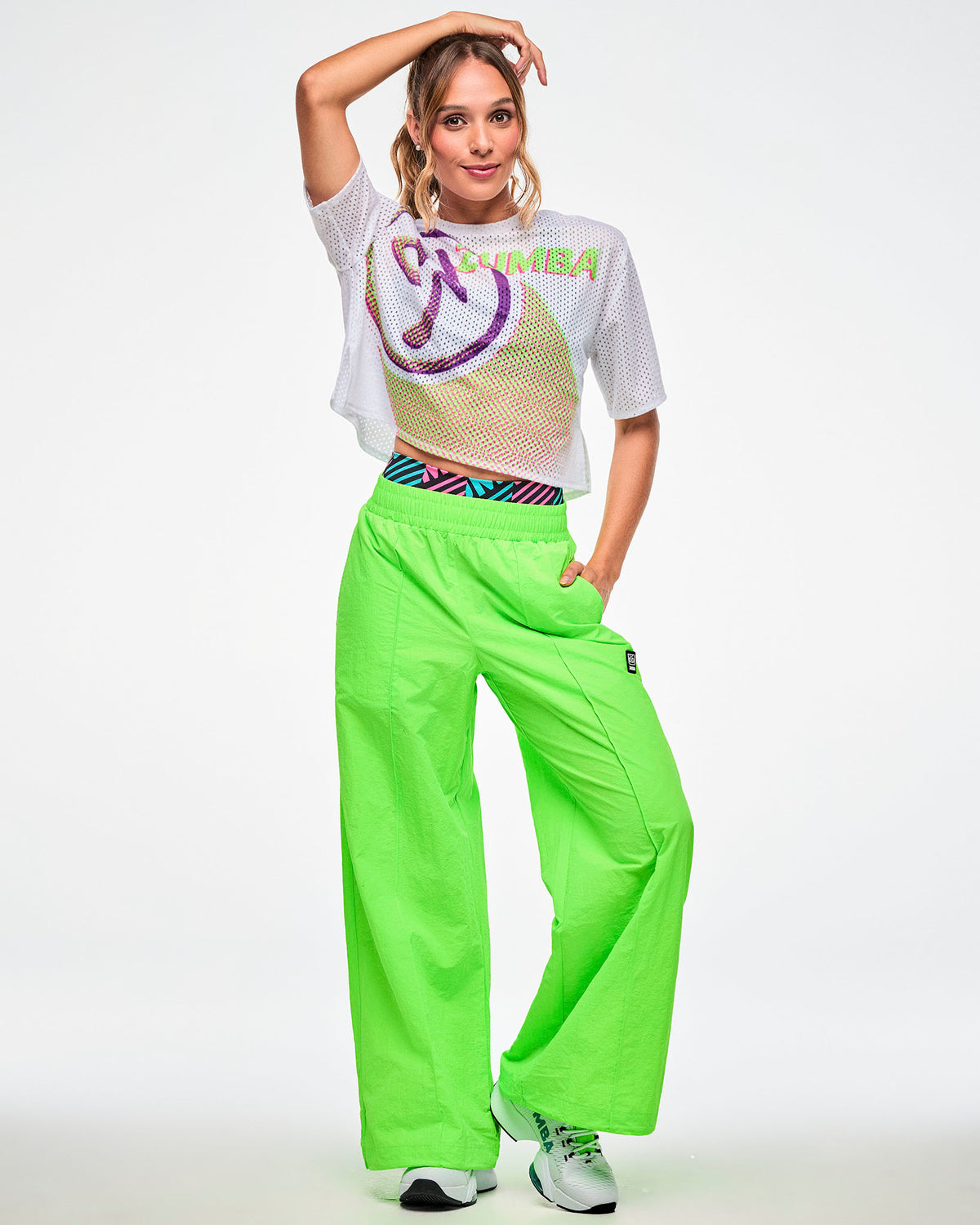 Full view of a female model wearing a white crop mesh top, paired with green pants.