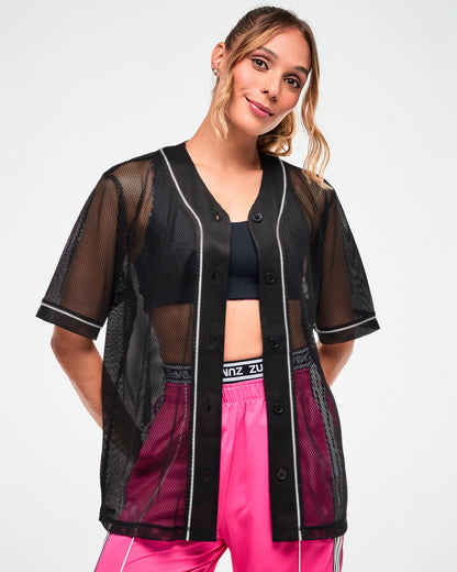 Close-up of a female model wearing a black mesh button-up baseball-style jersey with white piping, worn open over a black crop top and pink pants.