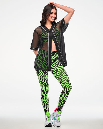Female model wearing a black mesh mostly unbuttoned baseball-style jersey over a green and black "ZUMBA" bra and paired with matching leggings and white shoes.
