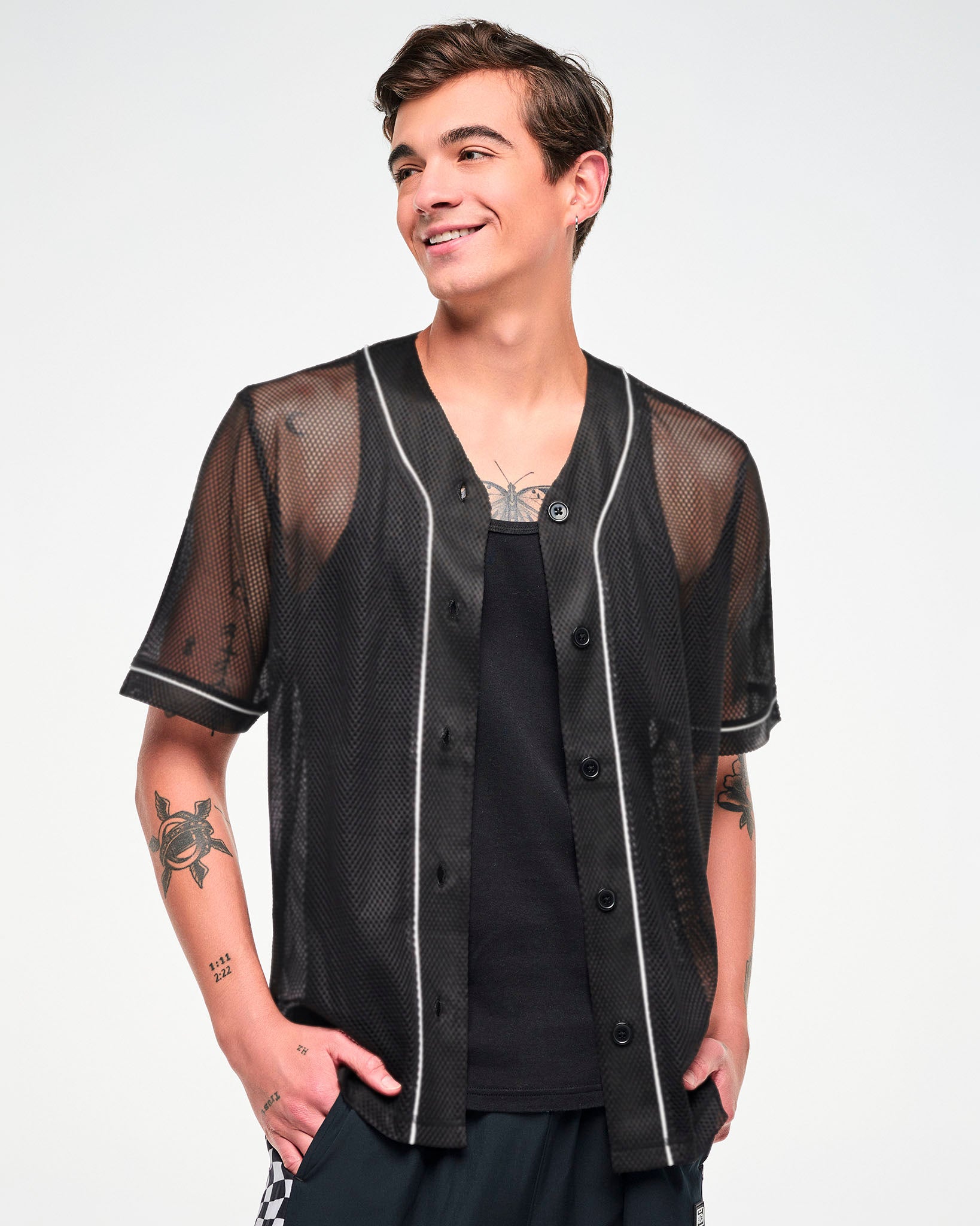 Male model wearing a black mesh baseball-style jersey with white piping, worn unbuttoned over a black tank top, paired with black pants.