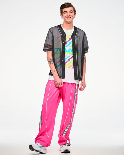 Male model wearing a black mesh baseball-style jersey unbuttoned, over a white "ZUMBA" graphic tee, paired with pink track pants and white sneakers.