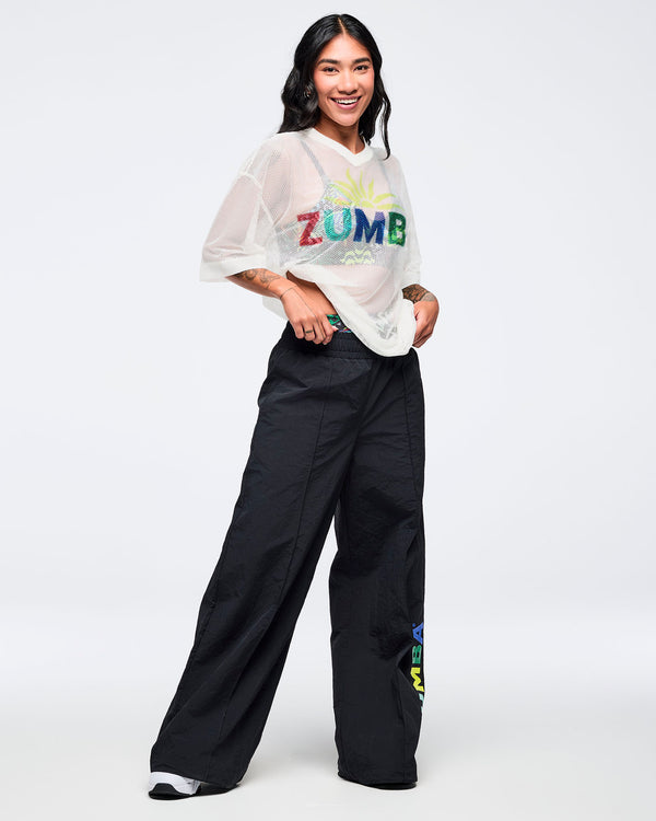 White mesh short-sleeve V-neck top with a multicolor “ZUMBA” chest graphic and relaxed fit, modeled by a female wearing black wide-leg pants with a multicolor “ZUMBA” leg print on a white studio background.