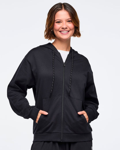 Front view of a female model wearing a black zip-up hoodie with hands in the pockets, layered over a white top, paired with black pants, against a white background.