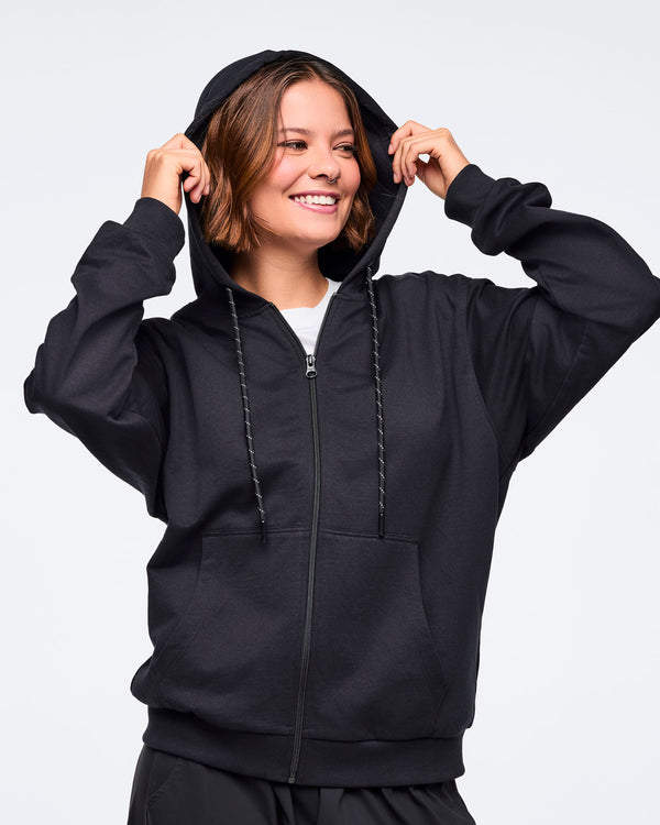 Front view of a female model pulling on the hood of a black zip-up hoodie, layered over a white top, paired with black pants, against a white background.