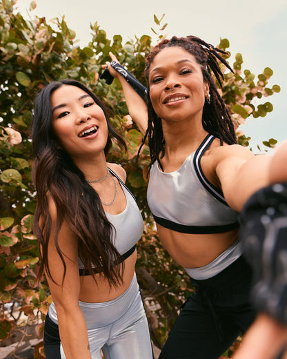Two female models pose for a selfie outdoors; one wears a silver sports bra with shiny silver leggings, and the other wears a silver sports bra with black jogger pants.