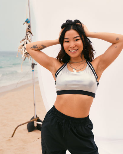 Female model wears the shiny silver cropped sports bra with relaxed black drawstring joggers, smiling with both hands in her hair in front of a white beach backdrop.
