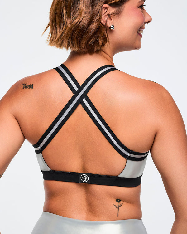 Metallic silver sports bra with black-and-silver crisscross back straps and a small center-back logo. Female model wears the glossy bra with matching metallic silver leggings featuring black side panels, shown in a close back view against a light gray background.