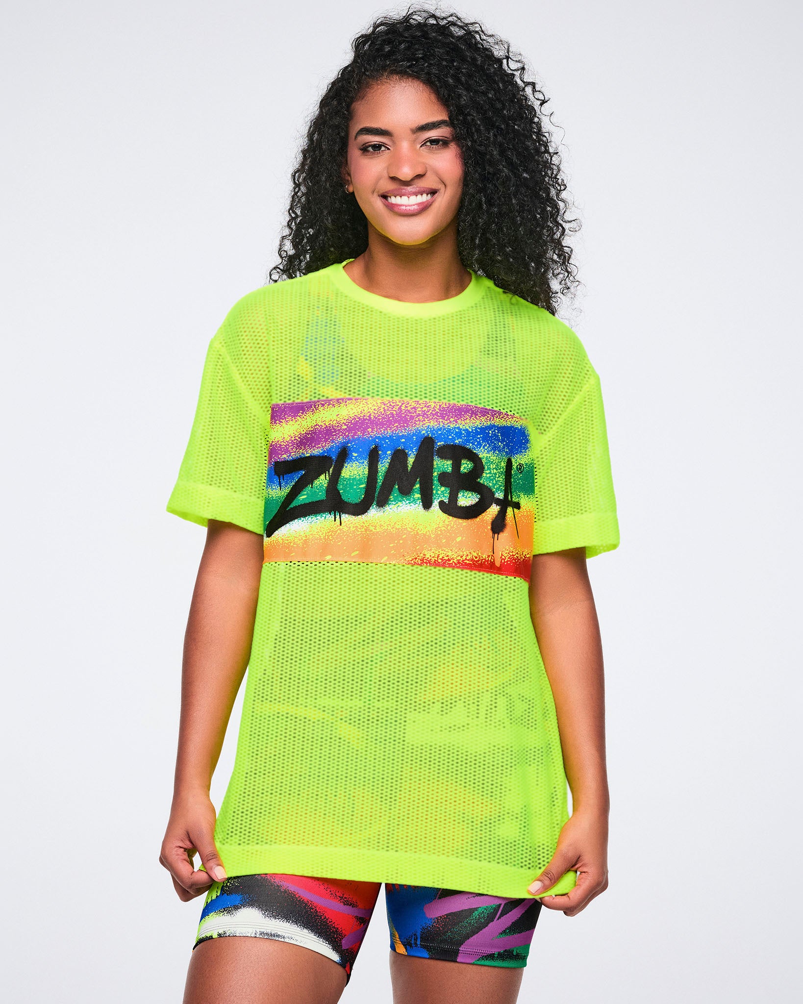 Female model wearing a neon yellow mesh Zumba shirt with colorful design on a white background.