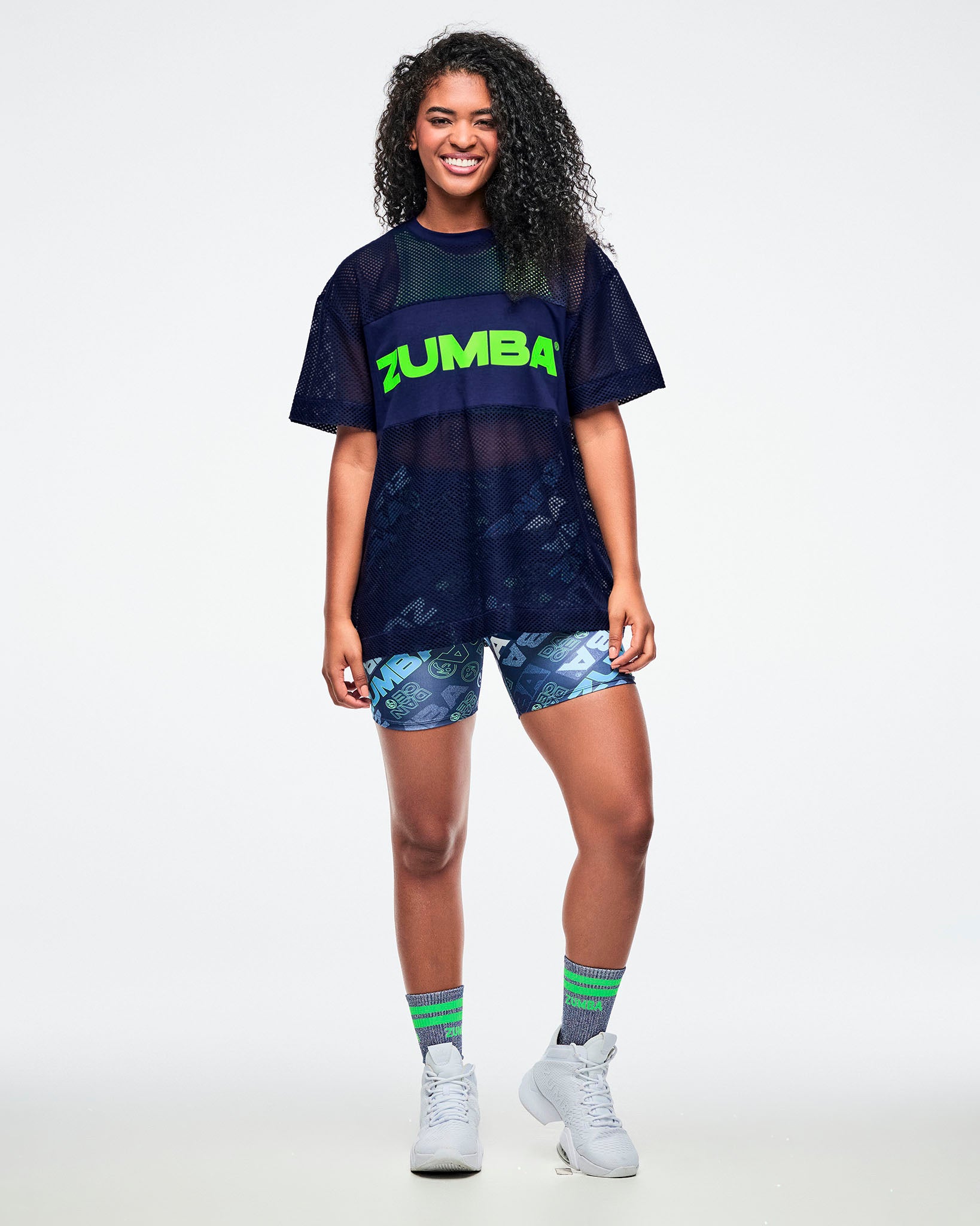 Female model in an indigo Zumba-branded mesh top over a green tank, paired with blue shorts, indigo socks, and white shoes.