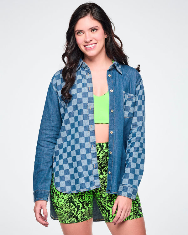 Front view of a female model wearing an open denim button-down with a checkerboard front panel, layered over a neon green bra top and paired with green snake-skin biker shorts.