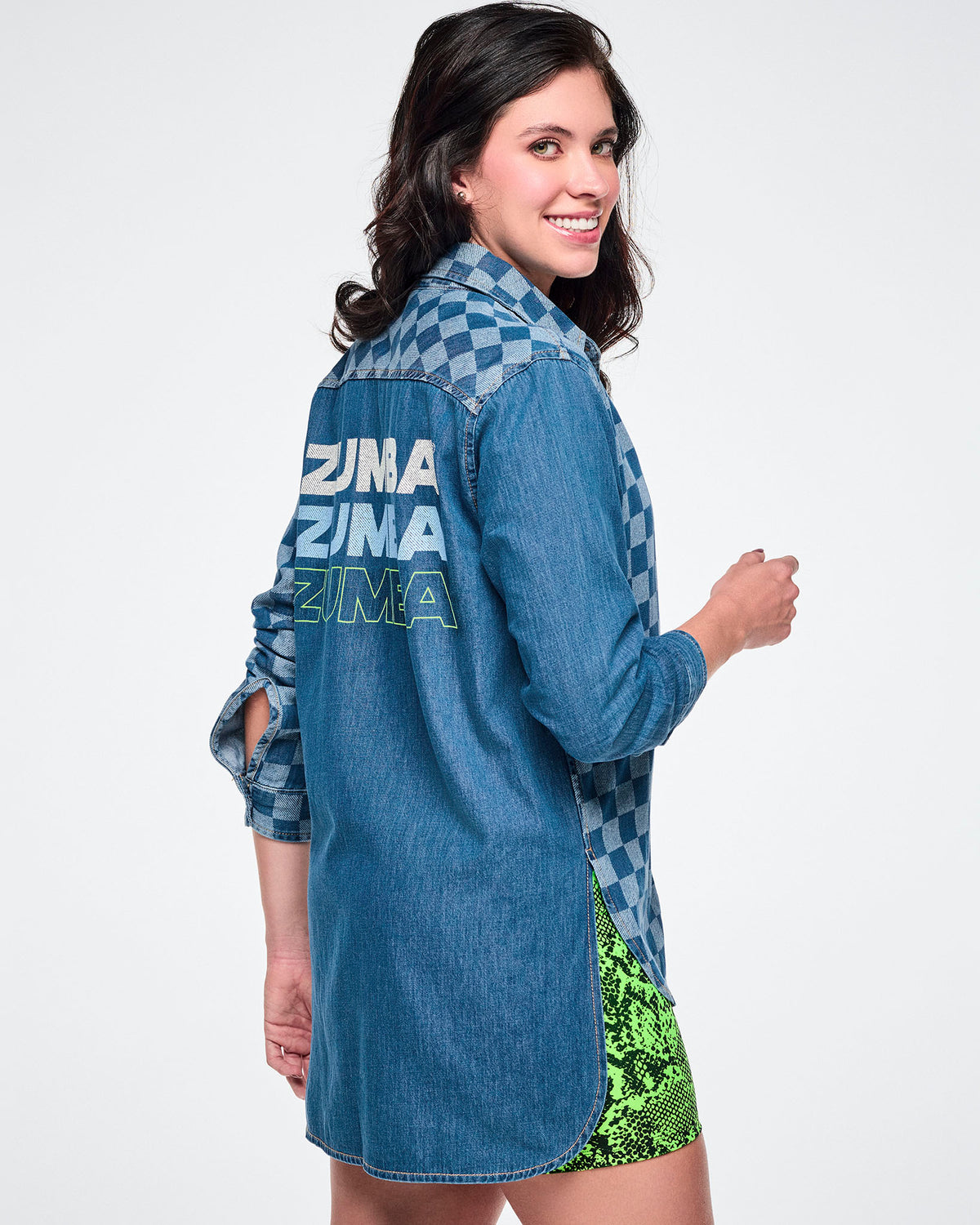 Side view of a female model wearing a denim button-down with a longer back hem and a checkerboard left sleeve, featuring "ZUMBA" repeated three times — white, then light blue, then outlined in neon green — paired with neon green snake-print shorts.