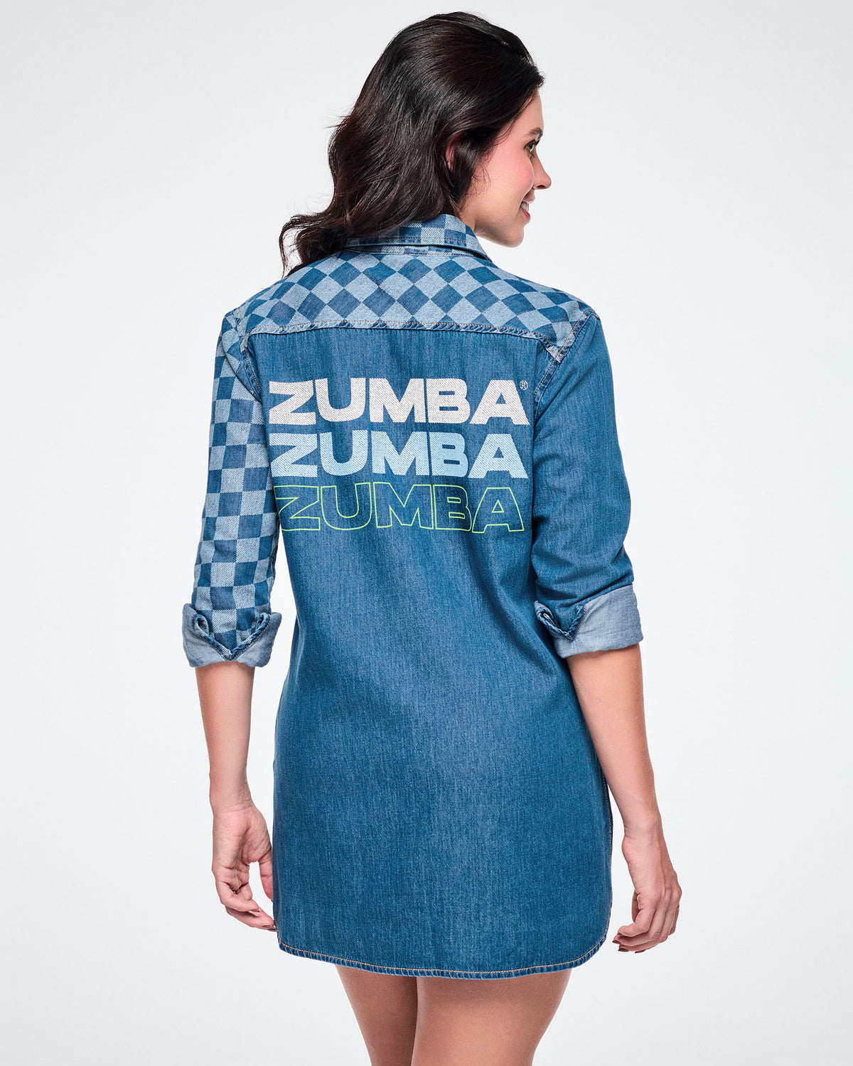 Back view of a female model wearing a denim button-down with a longer back hem and a checkerboard left sleeve, featuring "ZUMBA" repeated three times below the yoke — white, then light blue, then outlined in neon green.