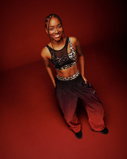A female model posing in a tan leopard print sports bra with asymmetrical mesh panel and gradient red and black pants, against a solid dark red studio background.