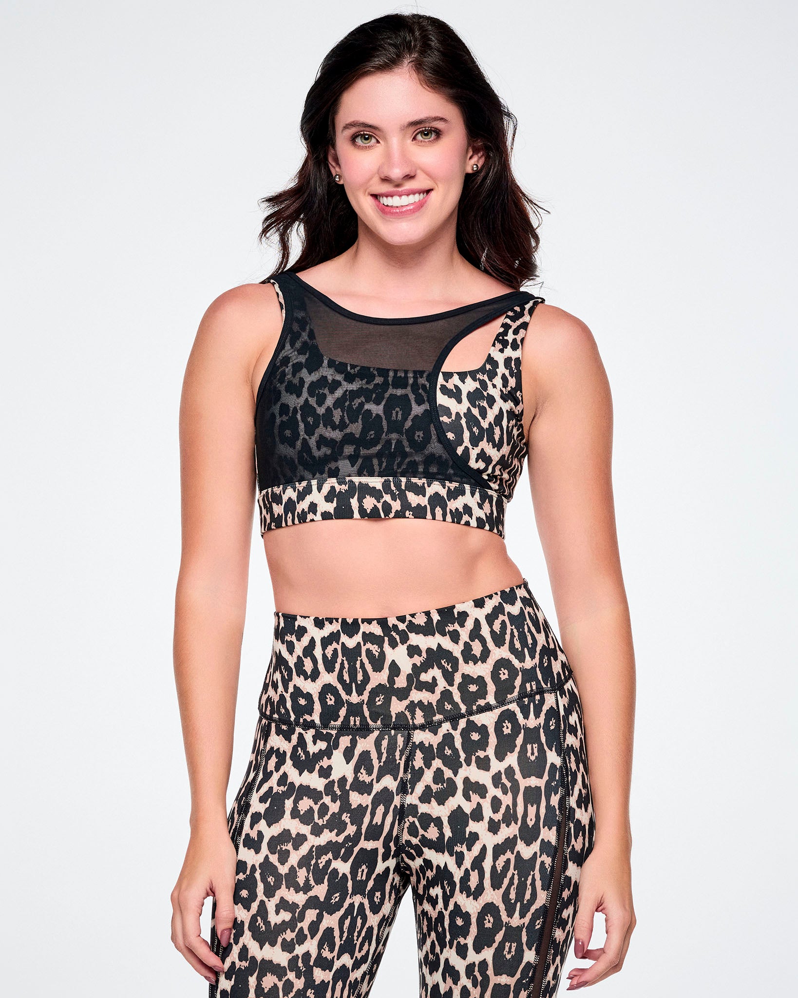 A female model wearing a sports bra with a tan leopard print and black asymmetrical mesh panel, paired with matching leggings.