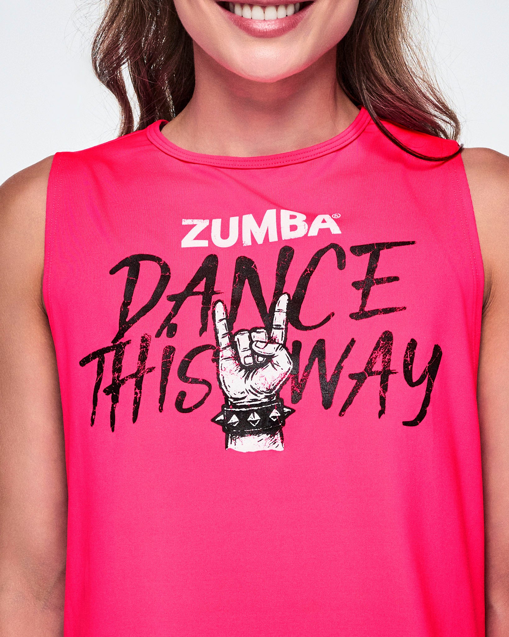Close-up of a female model wearing a bright pink sleeveless tank top with a Zumba graphic that reads 'Dance This Way' in black lettering, with a hand making a rock gesture and Zumba at the top in white.