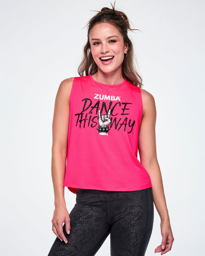 A smiling female model wearing a bright pink sleeveless tank top with a Zumba graphic that reads 'Dance This Way' in black lettering, with a hand making a rock gesture and Zumba at the top in white, paired with black leggings.