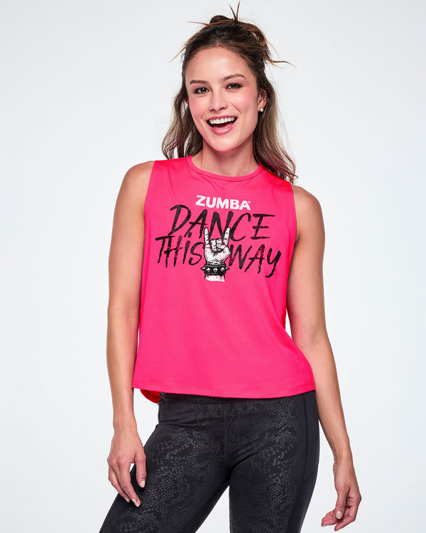 A smiling female model wearing a bright pink sleeveless tank top with a Zumba graphic that reads 'Dance This Way' in black lettering, with a hand making a rock gesture and Zumba at the top in white, paired with black leggings.