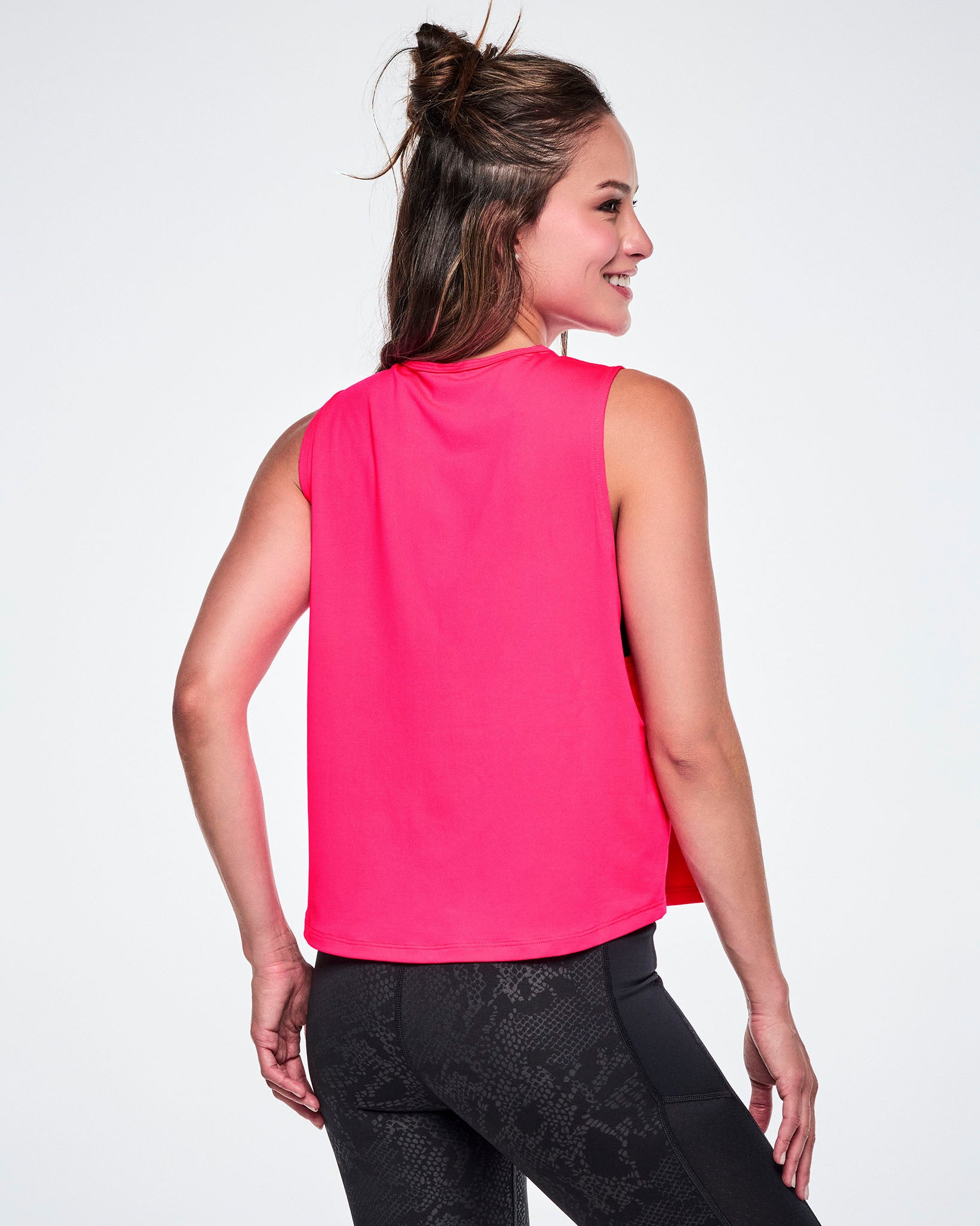 Back view of a female model wearing a bright pink sleeveless Zumba tank top, paired with black patterned leggings.