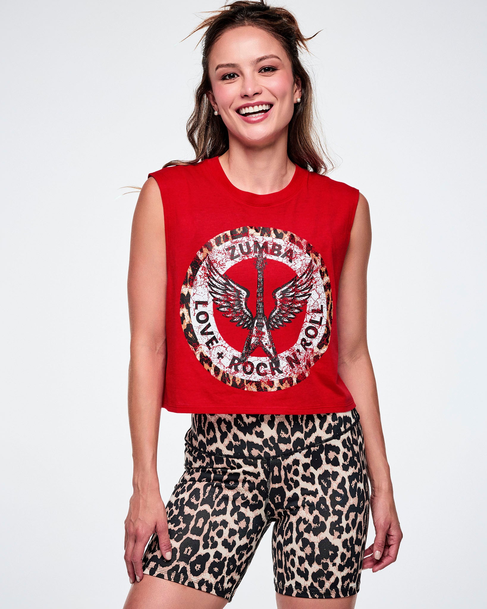 A female model wearing a red sleeveless crop tank with a large white, distressed, circular Zumba rock graphic on the front with a leopard print border. In the circle it says ZUMBA at the top and LOVE + ROCK N' ROLL at the bottom, with a white guitar and wings outlined in the middle. This is paired with tan leopard print biker shorts.