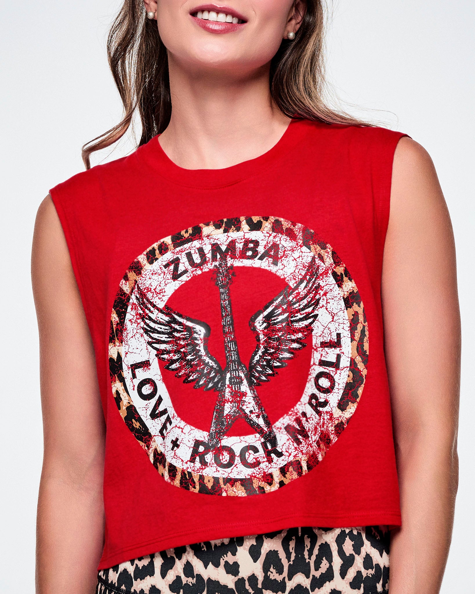 Close-up view of a female model wearing a red sleeveless crop tank with a large white, distressed, circular Zumba rock graphic on the front with a leopard print border. In the circle it says ZUMBA at the top and LOVE + ROCK N' ROLL at the bottom, with a white guitar and wings outlined in the middle.