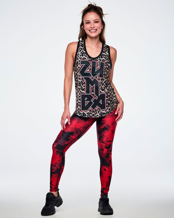 Full view of a female model wearing a basketball tank top with a black, beige, and white leopard print pattern, featuring large vertical 'ZUMBA' rock-like text in black with red and white outlines, paired with red and black leggings and black shoes.