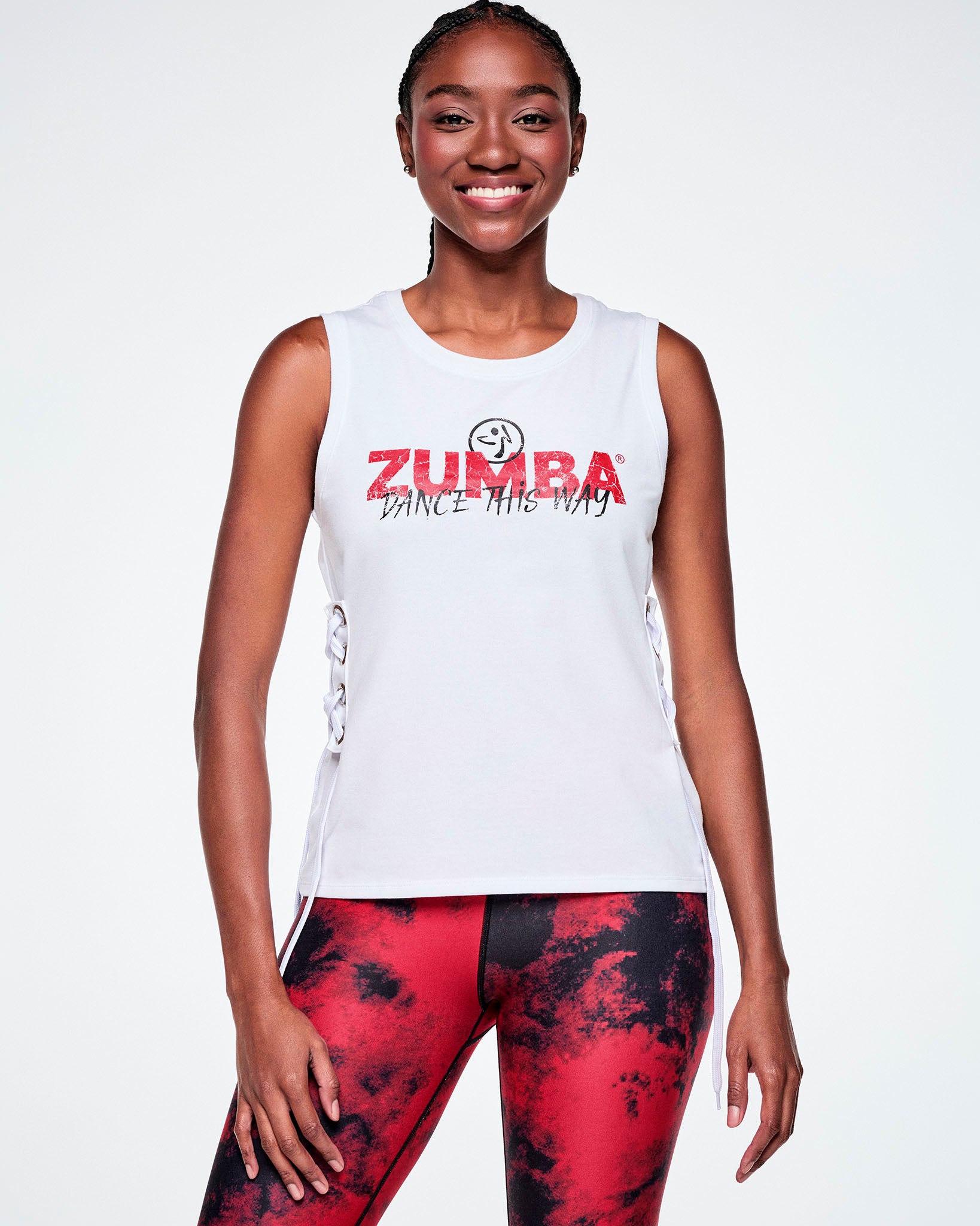Front view of a female model wearing a white sleeveless tank top with a red ZUMBA text and overlayed with a small black Zumbito on top and on the bottom, black cursive text saying "DANCE THIS WAY, all paired with red and black leggings.