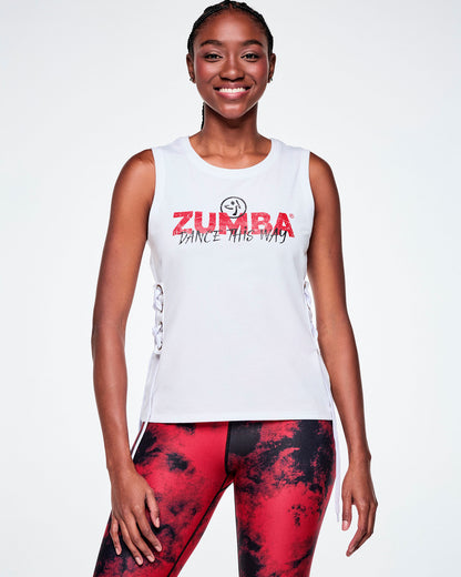 Front view of a female model wearing a white sleeveless tank top with a red ZUMBA text and overlayed with a small black Zumbito on top and on the bottom, black cursive text saying "DANCE THIS WAY, all paired with red and black leggings.
