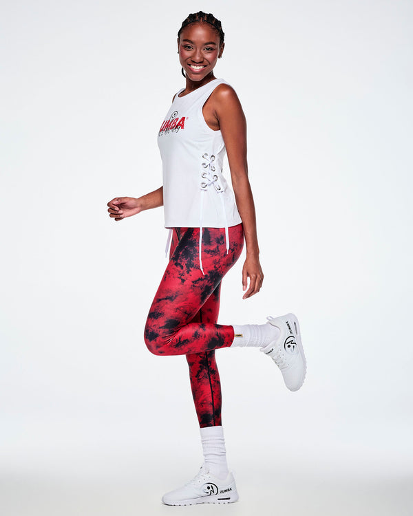 A female model wearing a sleeveless white tank top with red and black graphic lettering and side lace-up details, paired with red and black leggings and white shoes.