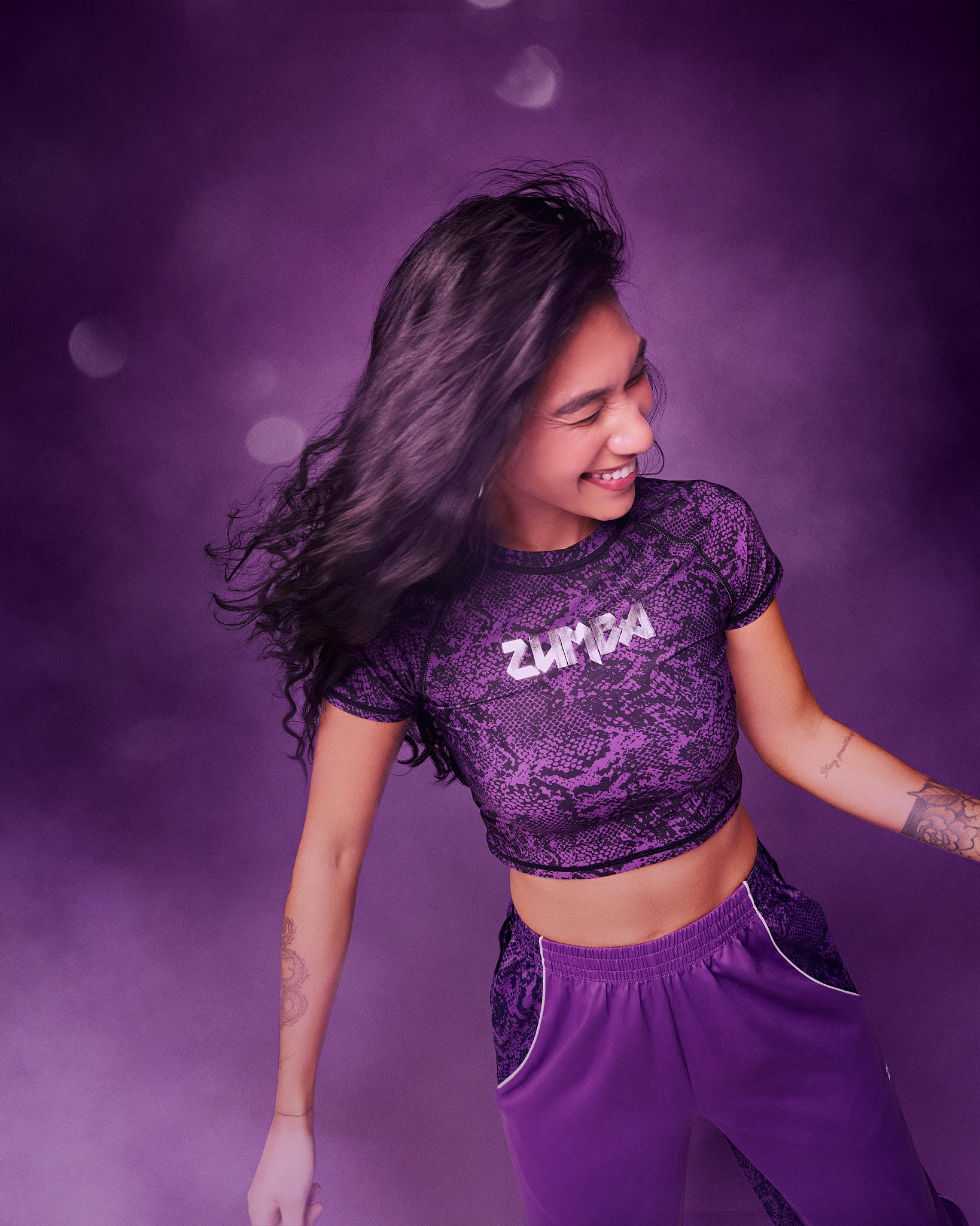 A female model, dancing wearing a purple Zumba crop top and matching purple pants, against a purple studio background with soft light bokeh.