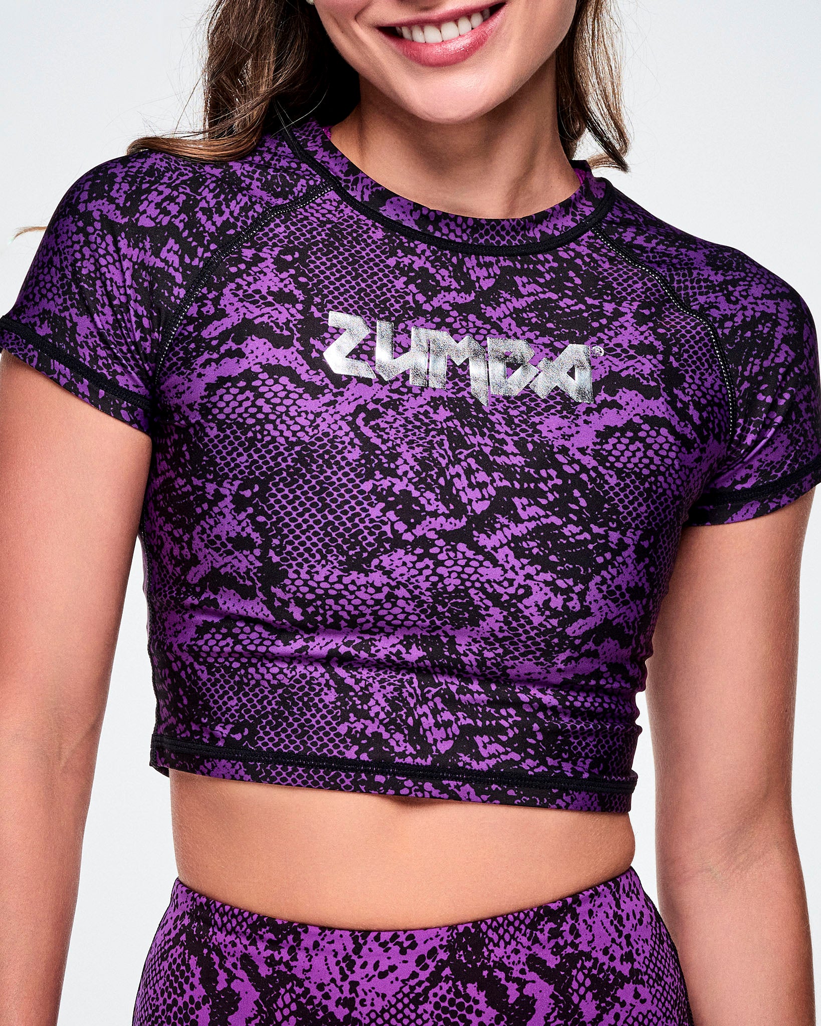 A close-up of a female model wearing a purple and black snakeskin-patterned short-sleeve crop top with the ZUMBA rock inspired text, printed in silver across the chest.