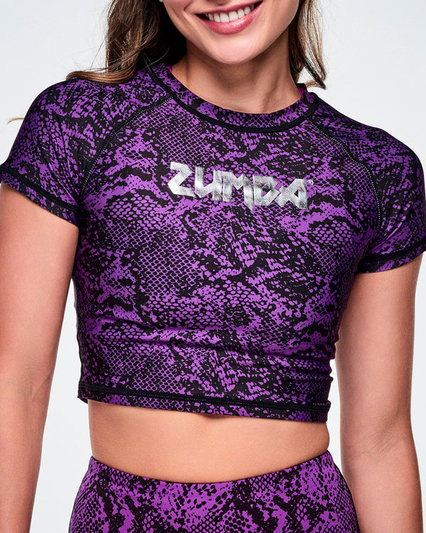A close-up of a female model wearing a purple and black snakeskin-patterned short-sleeve crop top with the ZUMBA rock inspired text, printed in silver across the chest.