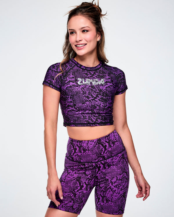 Full view of a female model wearing a purple and black snakeskin-patterned short-sleeve crop top with the ZUMBA rock inspired text, printed in silver across the chest and paired with matching biker shorts.
