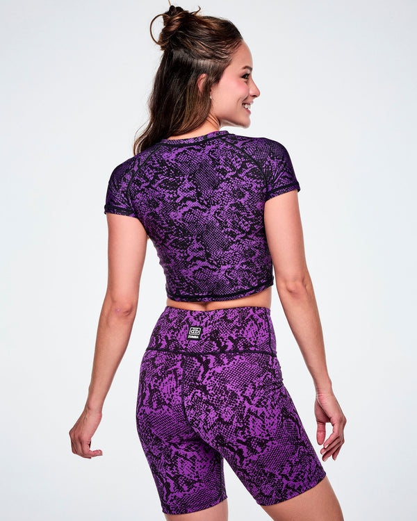 Back view of a female model wearing a purple and black snakeskin-patterned short-sleeve crop top and paired with matching biker shorts.