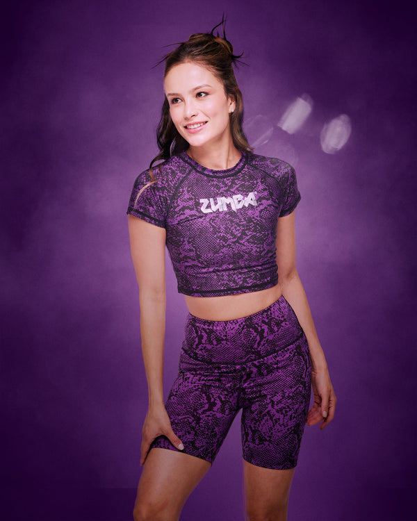 A female model wearing a matching purple and black snakeskin-patterned activewear set consisting of a short-sleeve crop top with 'ZUMBA' printed on the chest, in silver and matching high-waisted bike shorts, set against a smoky purple studio background.