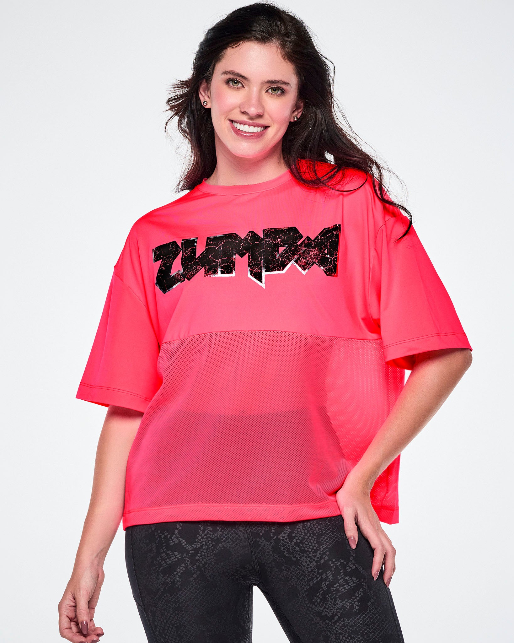 A female model wearing a bright pink short-sleeve top with mesh bottom panels and black and silver distressed  'ZUMBA' rock-like text across the chest, paired with black leggings.