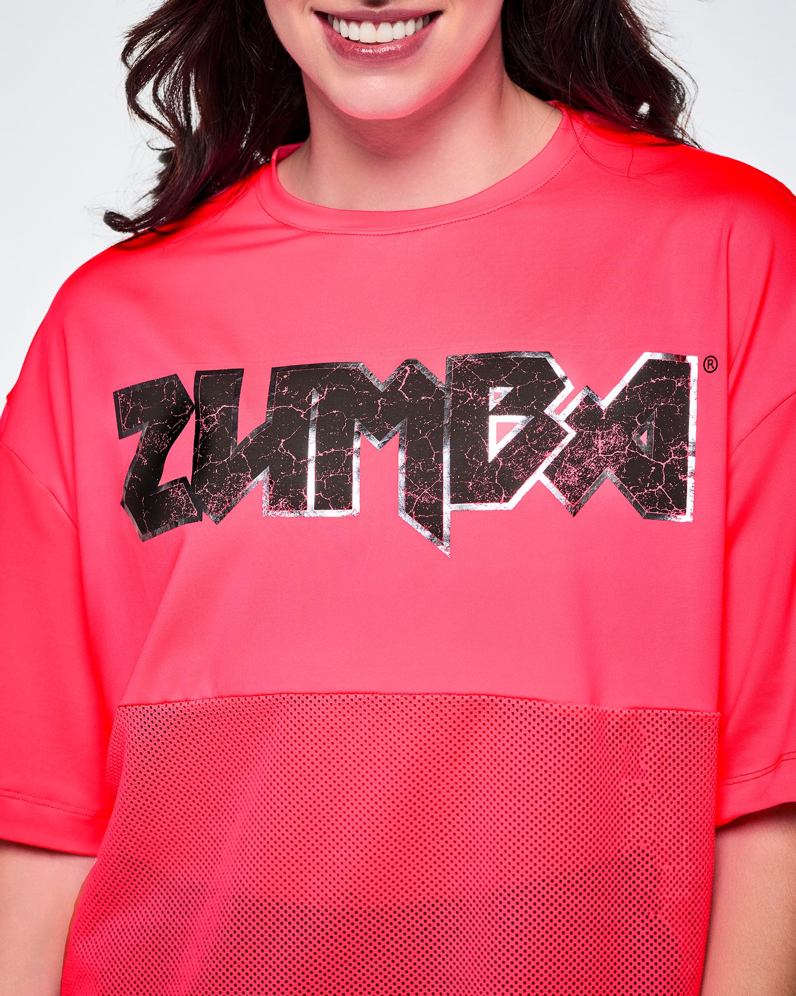 A close-up of a female model wearing a bright pink short-sleeve top with mesh bottom panels and black and silver distressed 'ZUMBA' rock-like text across the chest.