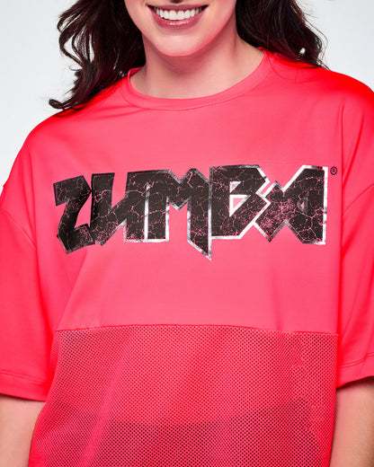 A close-up of a female model wearing a bright pink short-sleeve top with mesh bottom panels and black and silver distressed 'ZUMBA' rock-like text across the chest.
