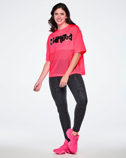 Full view of a female model wearing a bright pink short-sleeve top with mesh bottom panels and black and silver distressed 'ZUMBA' rock-like text across the chest, paired with black leggings and pink shoes.