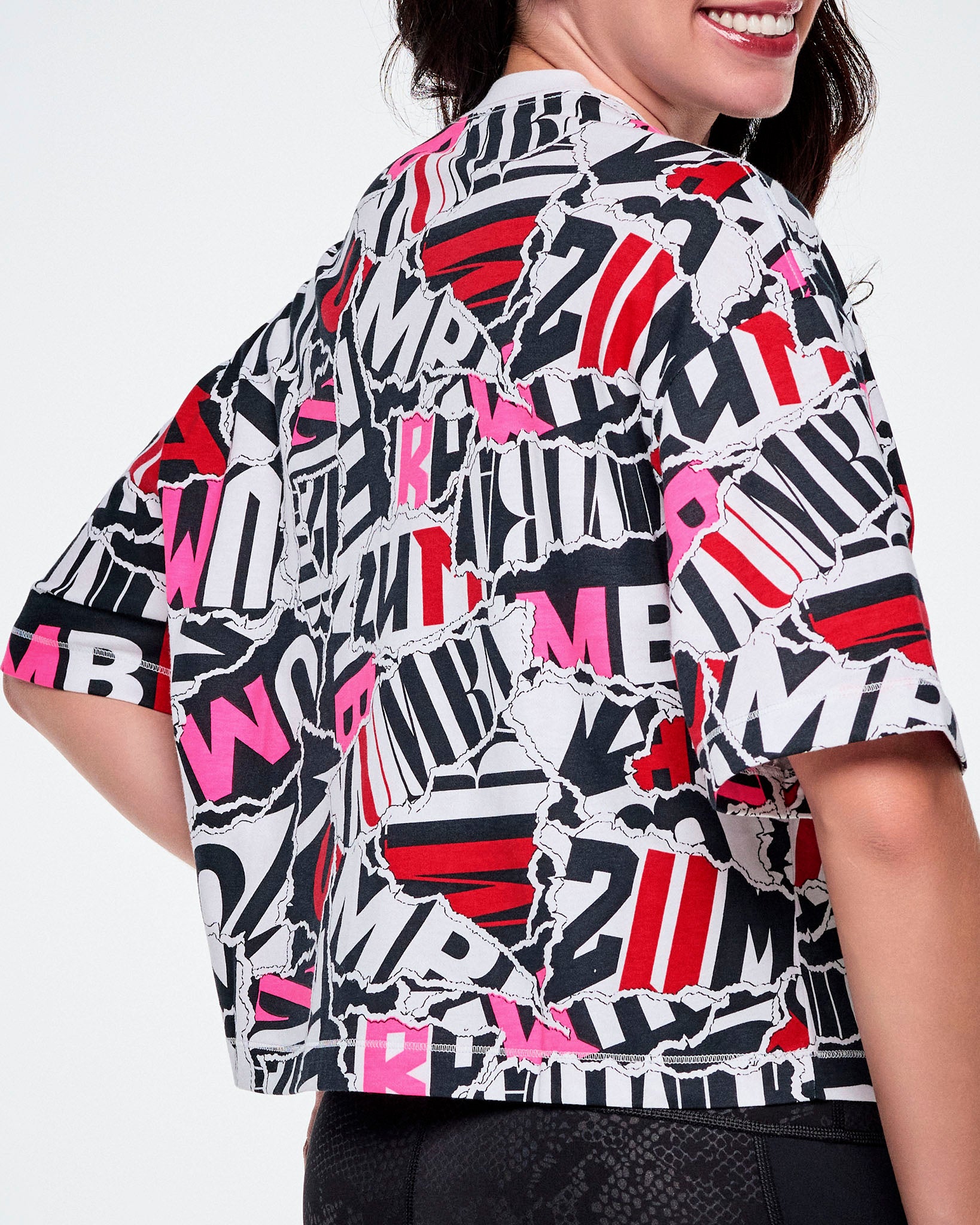 A close-up back view of a female model wearing a short-sleeve crop top with a bold abstract ZUMBA text pattern print in black, white, red, and pink.